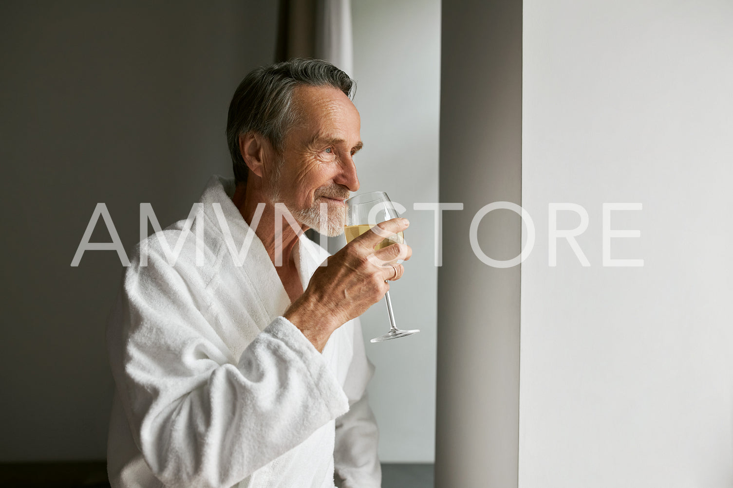 Senior man in bathrobe smell wine looking at window in hotel room	