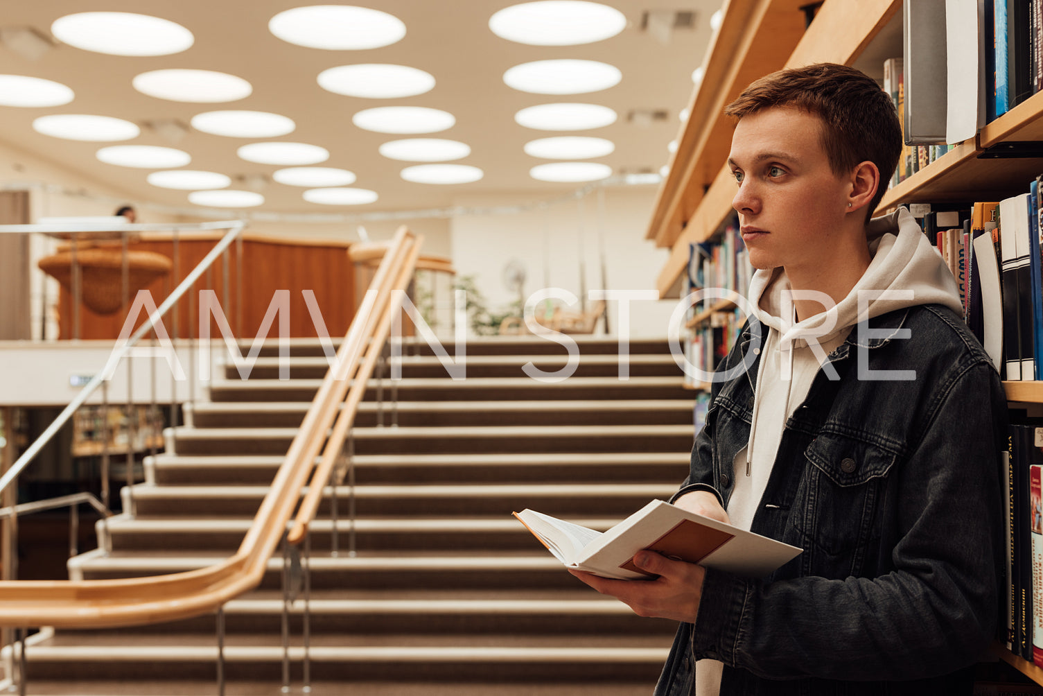 Side view of male student in library holding a book. University student standing at bookshelf.