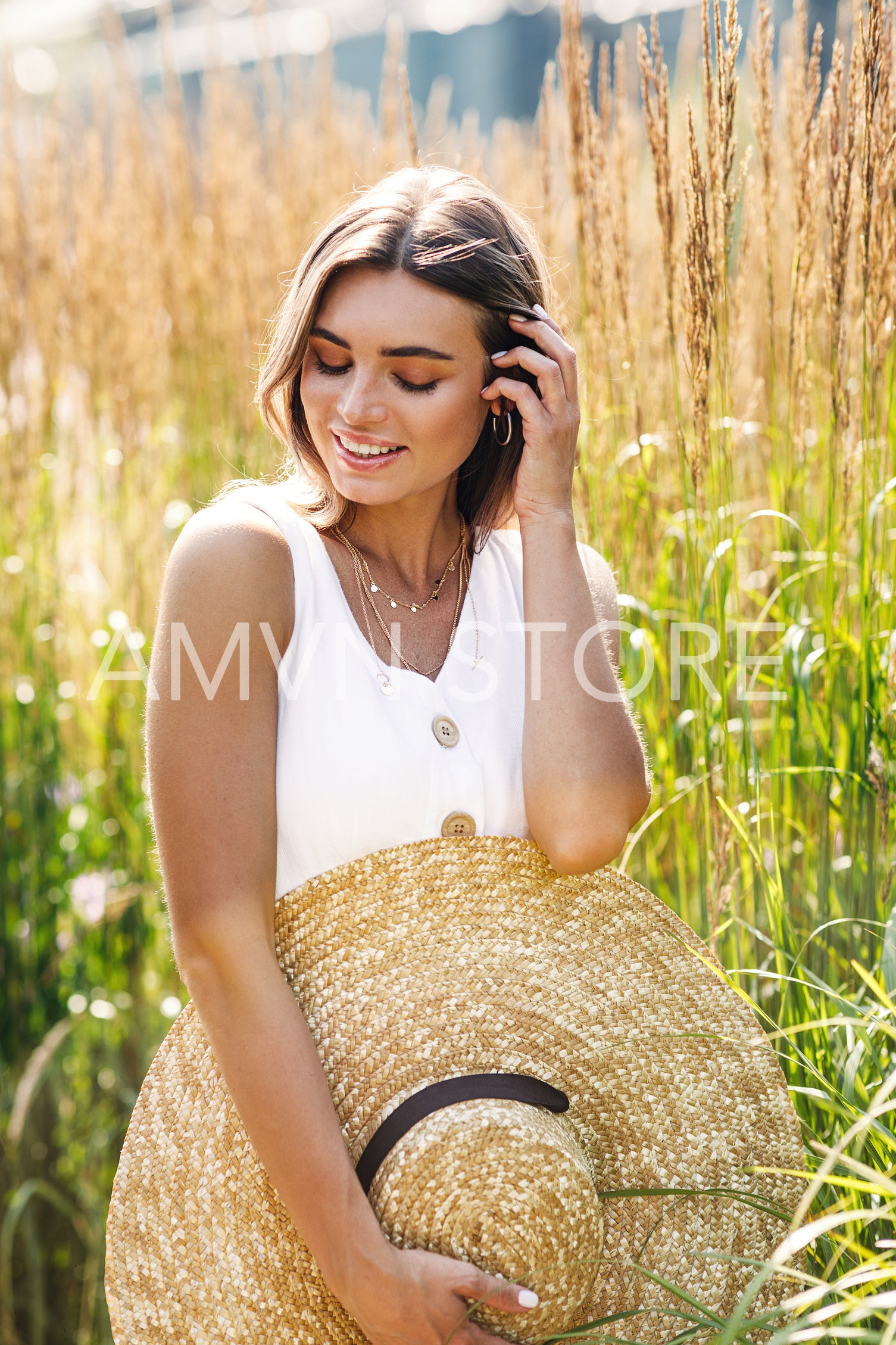 Young smiling woman holding her big straw hat and looking down while standing on the field	