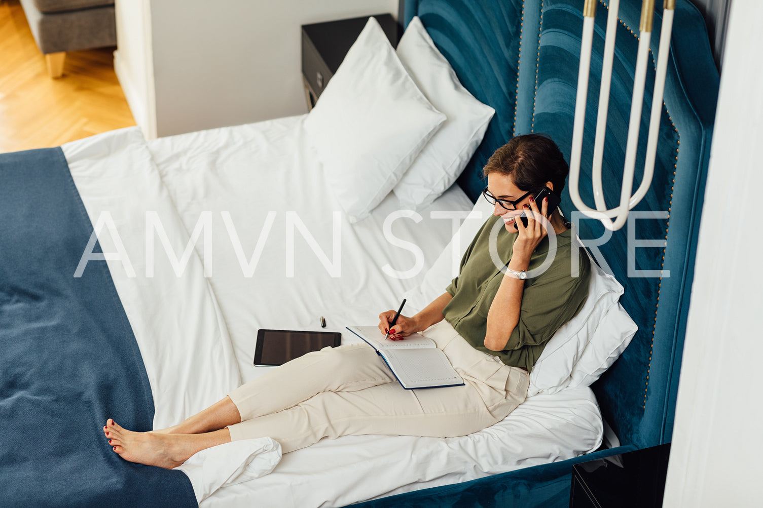 High angle view of woman in formal clothes lying on bed talking over mobile phone with a notebook on her hips	