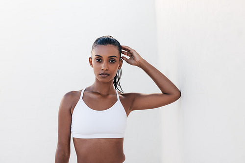 Woman leaning on a wall after fitness session in a white outdoor studio. Portrait of a young female in sportswear posing outdoors looking at the camera.