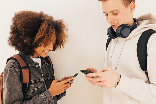 Two college students standing together at wall looking at their smartphones