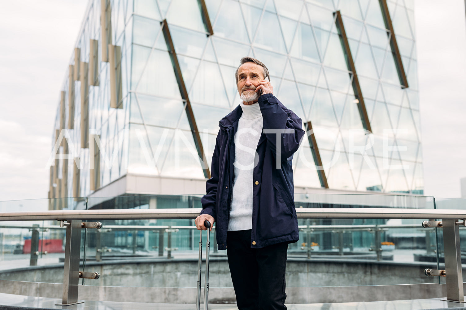 Mature man standing in front of office building with suitcase and talking on cell phone	