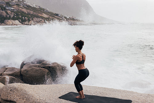 Woman practicing yoga while standing on stone against big waves and splashes. Female meditating at oceanside.