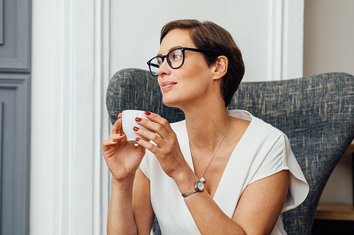 Businesswoman sitting on armchair in her apartment and holding a cup of coffee