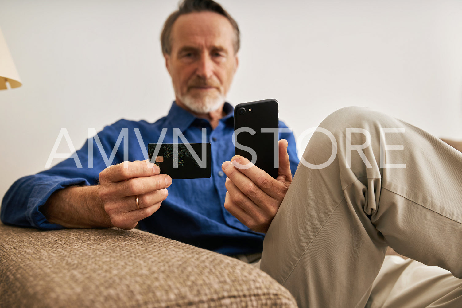 Senior man paying online. Male in casual clothes sitting on sofa holding a credit card and smartphone.