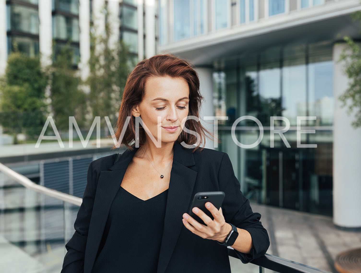 Middle-aged businesswoman looking at her mobile phone while leaning a railing against an office building