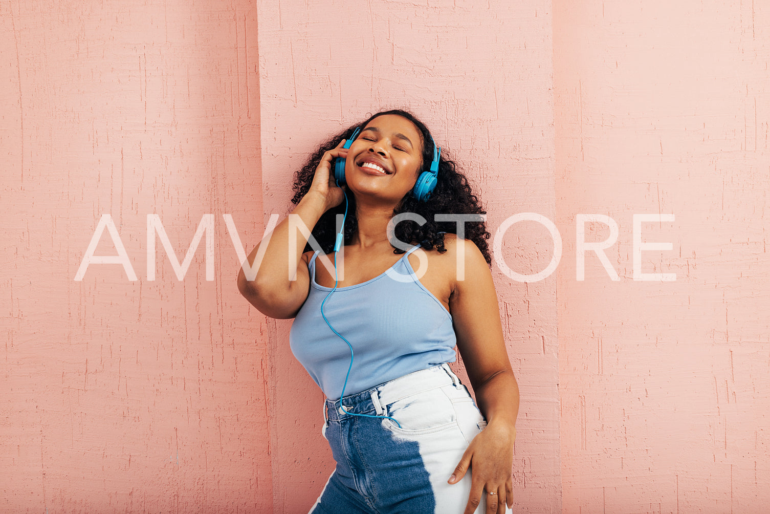 Young woman enjoying music while leaning pink wall. Female wearing blue headphones with closed eyes.