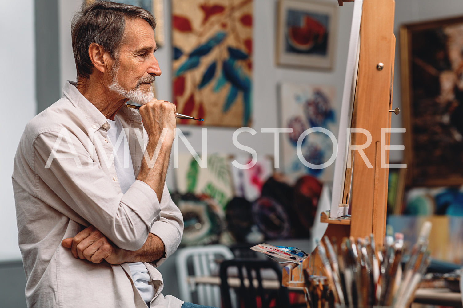 Side view of a thoughtful artist holding paintbrush while sitting in his studio. Male painter looking on canvas.	