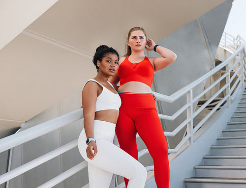 Two plus-size female athletes standing together and looking away while standing on stairs. Two women of different races in fitness attire relaxing outdoors.