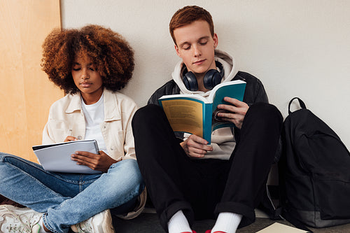 Two university students sitting at wall and reading. Classmates sitting together in corridor with books.