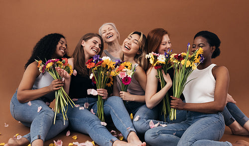 Six women of different ages and body types holding bouquets while sitting in studio against a brown background