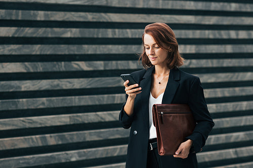 Smiling businesswoman holding a smartphone and leather folder standing outdoors. Middle-aged female with ginger hair typing on smartphone.