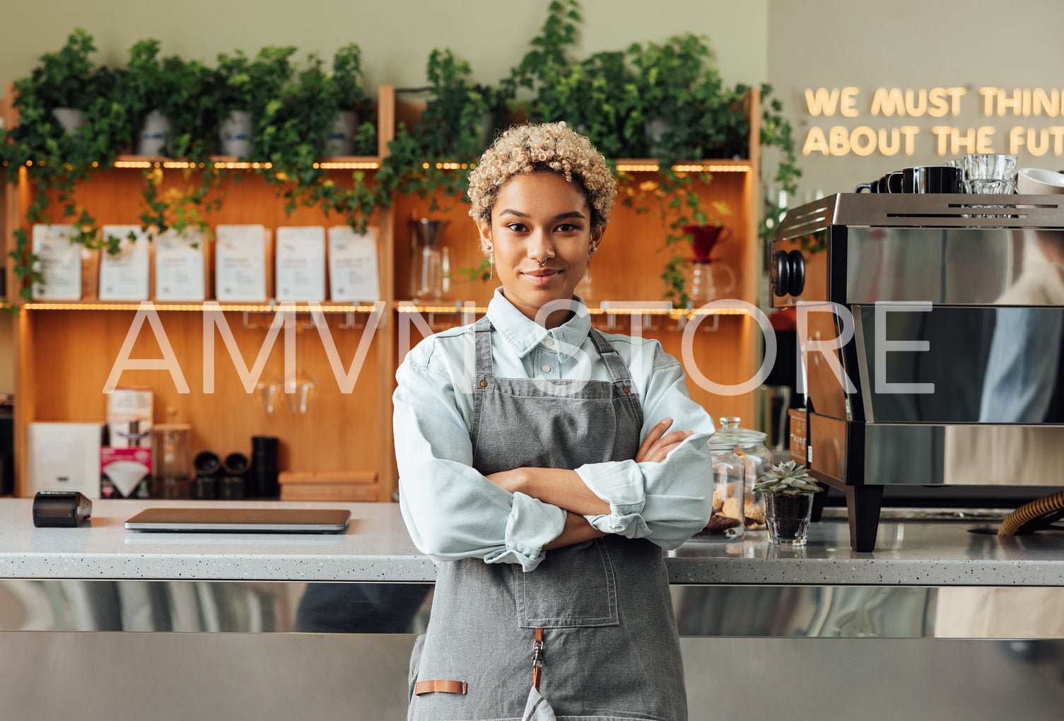 Portrait of a beautiful barista in an apron. Smiling coffee shop owner with crossed arms leaning at counter.