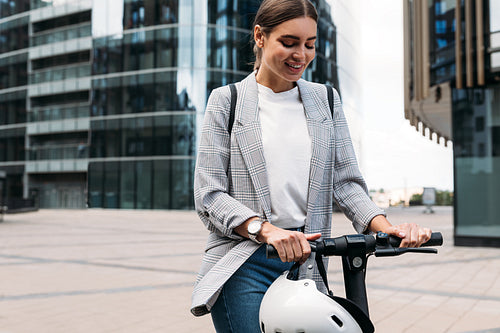 Smiling blond woman holding a handlebar of an e-scooter. Female in formal wear standing on electric push scooter ready to ride.