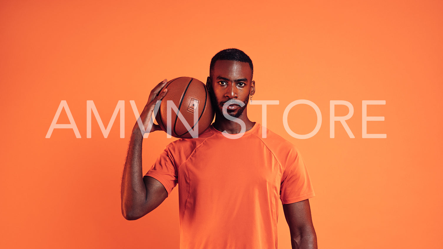 Serious basketball player looking at camera. Portrait of a male with basket ball on his shoulder against an orange background.