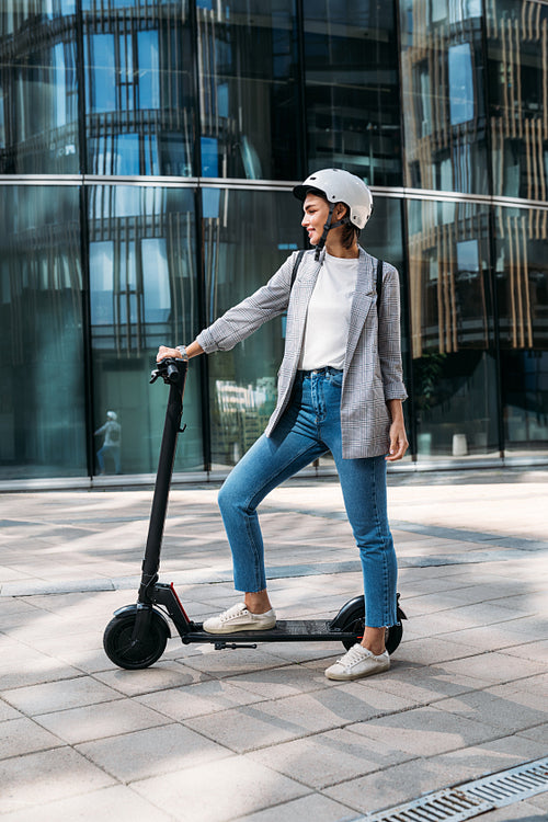 Side view young woman standing on electrical scooter near an office building. Female in helmet ready for a ride on scooter.