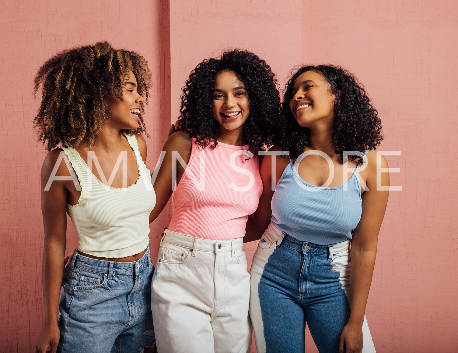 Three cheerful women with curly hair having fun together while standing at a pink wall