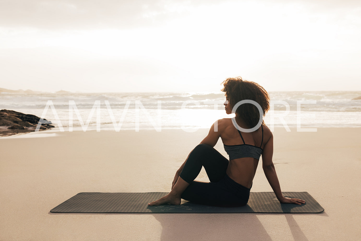 Side view of woman sitting on mat. Female meditating on seashore at sunset.