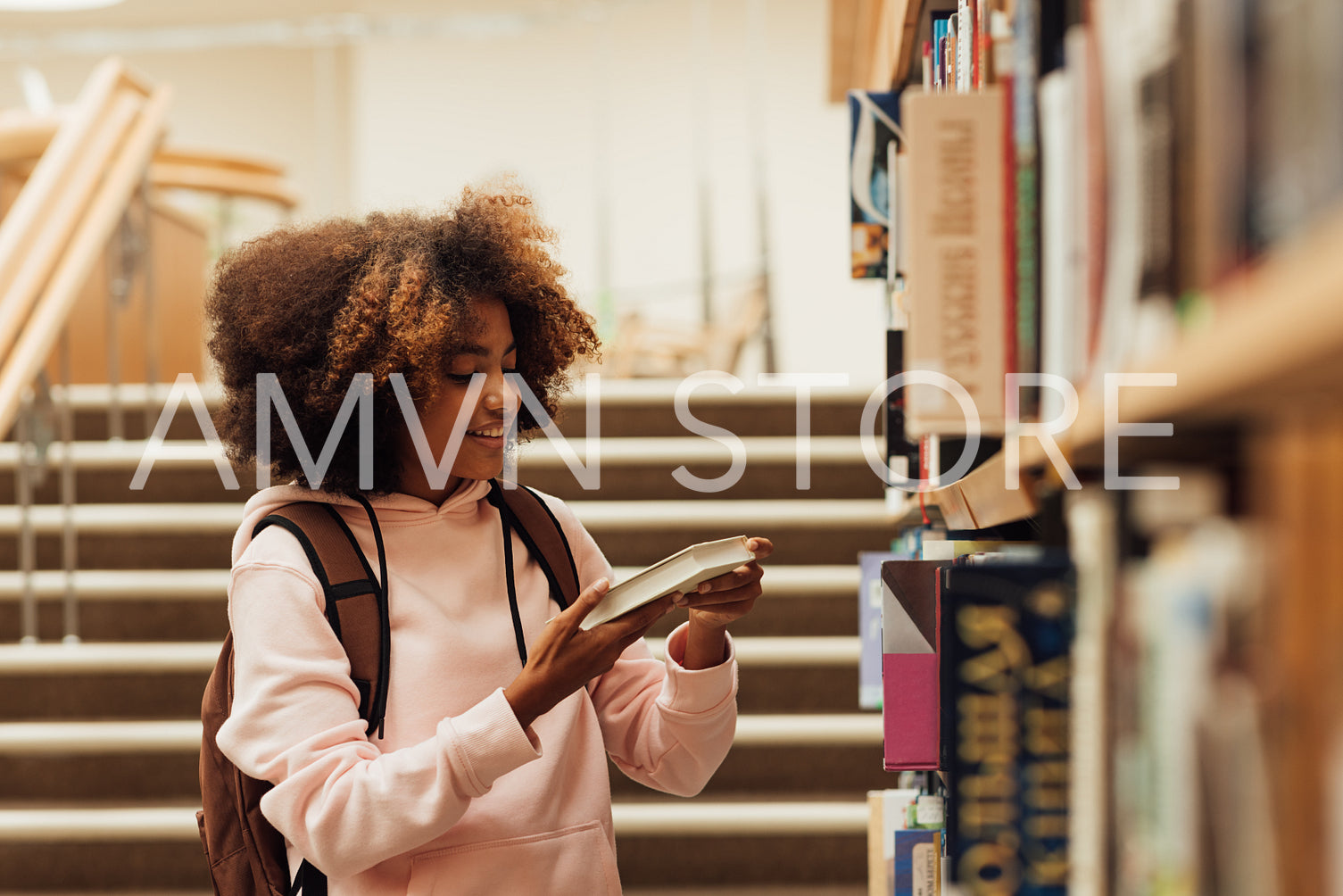 Smiling girl with curly hair holding a book near bookshelf in school library