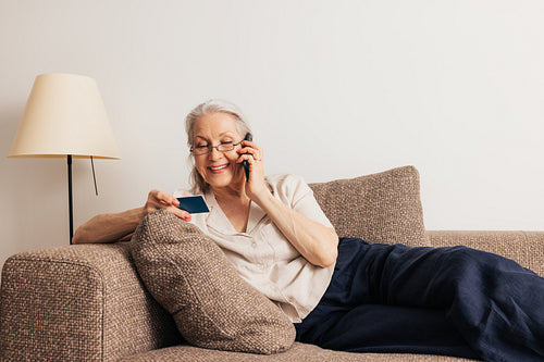 Senior woman looking at credit card talking on a mobile phone. Aged female making online shopping while lying on a sofa.