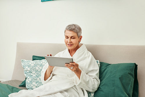 Smiling mature woman in bathrobe relaxing on a bed holding a digital tablet