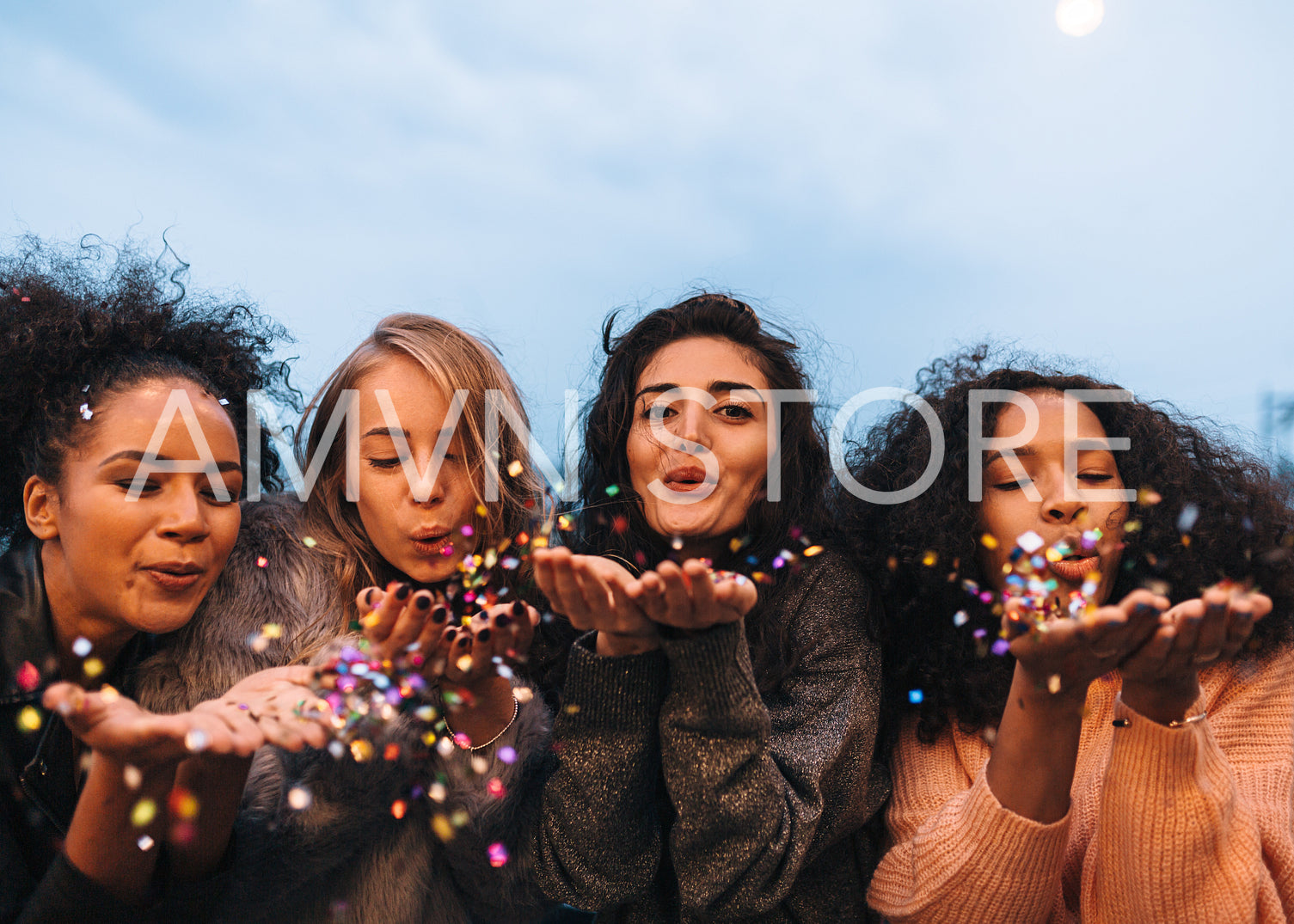 Group of four diverse females blowing colorful confetti from their hands at evening