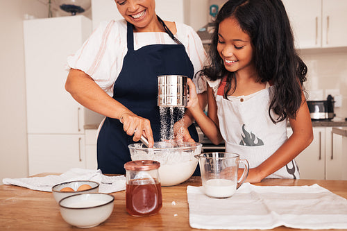 Senior woman and granddaughter sifting flour at a table in kitchen. Girl and her grandma making dough.