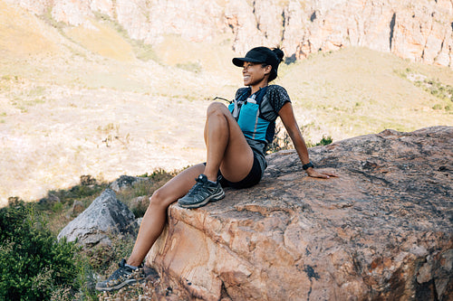 Woman in sports wear sitting on big stone taking a break during mountain hike. Female enjoying the view in valley.