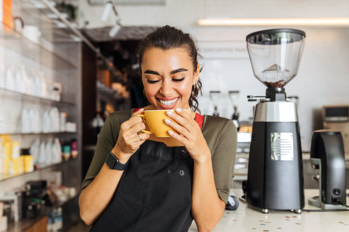 Happy barista standing inside coffee shop with closed eyes and holding a mug. Cheerful woman in apron standing at cafe counter.