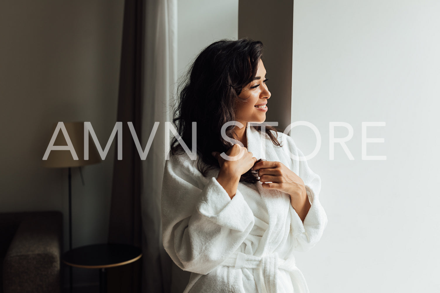 Side view of a brunette woman in a white bathrobe standing in living room and looking at a window	