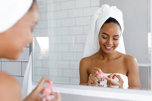 Happy young woman applying face cream in bathroom, standing in front of a mirror