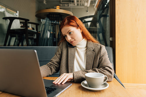 Woman working as a freelancer in a cafe. Female in formal wear typing on laptop and talk on a mobile phone.