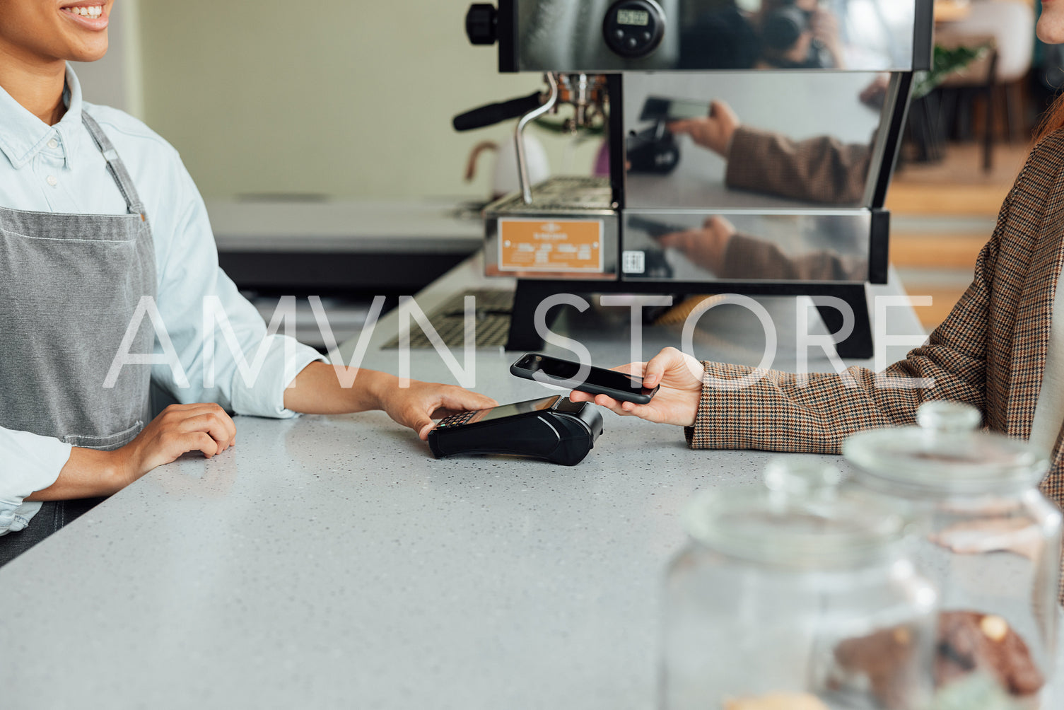 Side view of a barista at counter receiving payment from a customer in a cafe. Unrecognizable female paying by mobile phone using NFC at the counter.