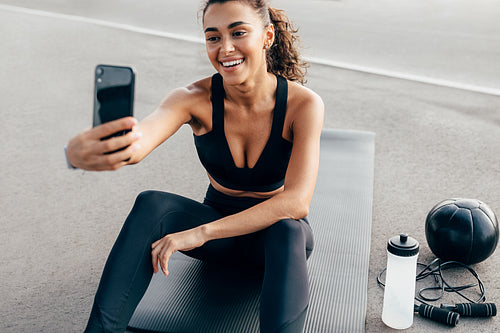 Happy woman with mobile phone sitting on a mat outdoors and live streaming during work out