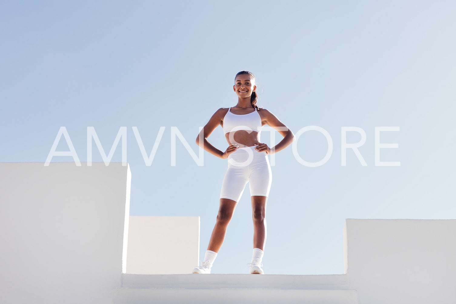 Slim smiling woman in white fitness attire posing on a top of stairs case. Full-length fit female with hands on her hips looking at the camera while standing on a top.