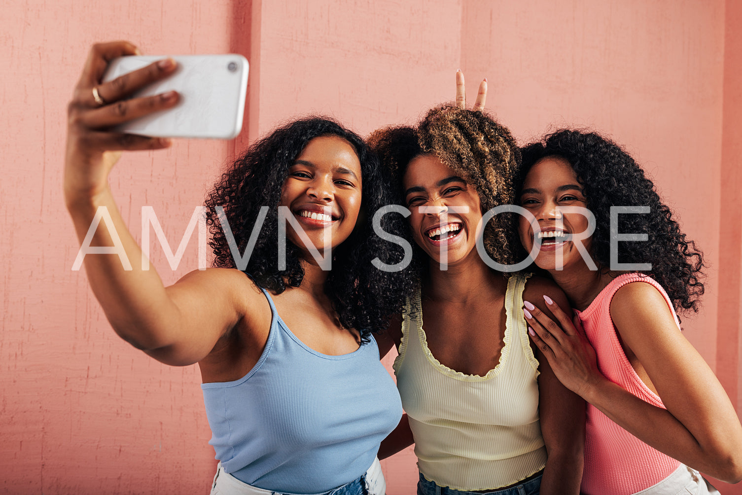 Three women with curly hair laughing together while taking selfie against a pink wall