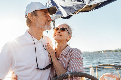 Happy mature woman looking on her husband while he steering a yacht. Affectionate couple enjoying a vacation on private sailboat at sunny day.