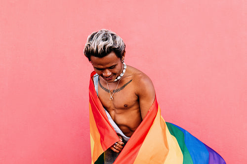 Young bare chested guy having fun while standing against pink wall with LGBT flag