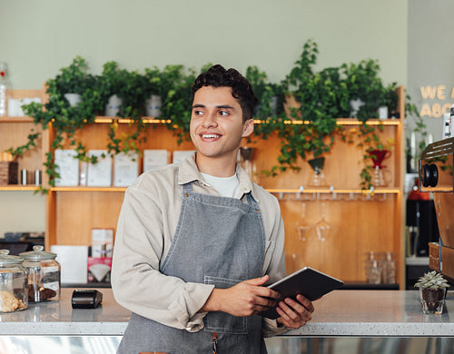 Male coffee shop owner in apron leaning counter. Middle eastern barista holding digital tablet looking away.