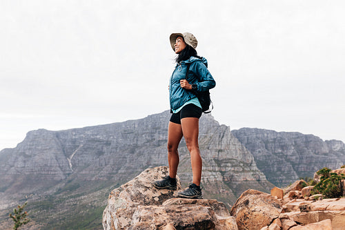 Smiling woman hiker wearing a hat enjoying the view while standing on an edge. Young female with a backpack on a hike.