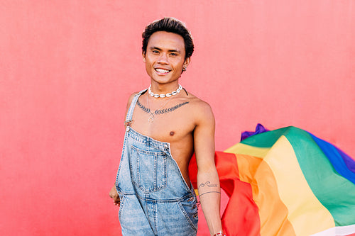 Smiling guy holding a rainbow LGBT flag while standing against a pink wall outdoors