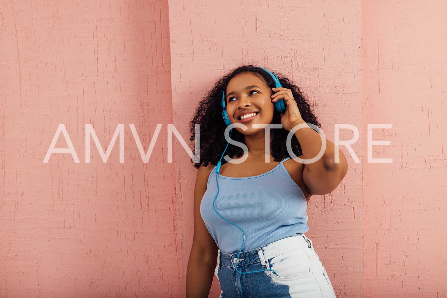 Happy woman wearing blue headphones leaning a pink wall 