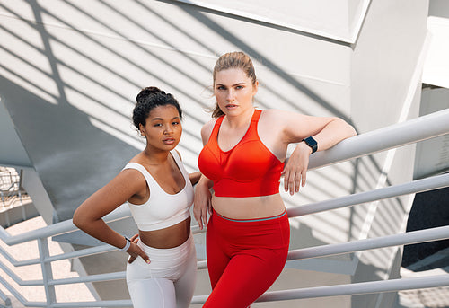 Portrait of two plus-size females in sportswear standing together. Two women in fitness attire standing on the stairs of a bridge and looking at camera.