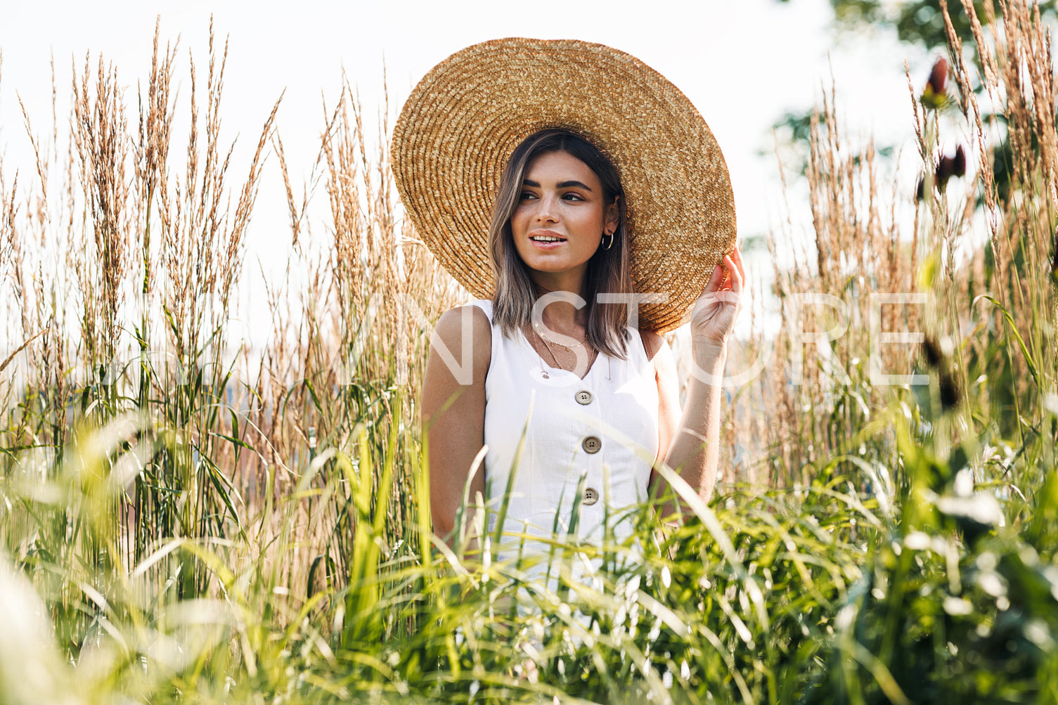 Young happy woman in big straw hat standing on the field and looking away. Stylish female walking outdoors.	