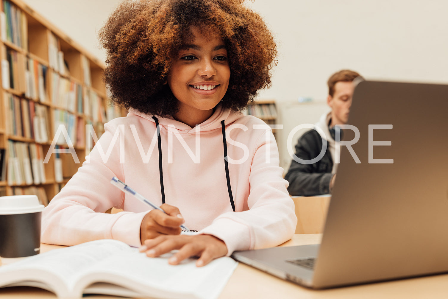 Smiling girl writing in a book while sitting at desk with laptop. Female student studying at college library.