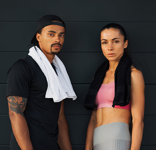 Two young athletes at a black wall. Fitness couple with towels relaxing at a wall.