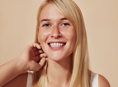 Young happy female with blond hair looking at camera in a studio. Beautiful smiling woman with freckles adjusting her hair over a beige backdrop.