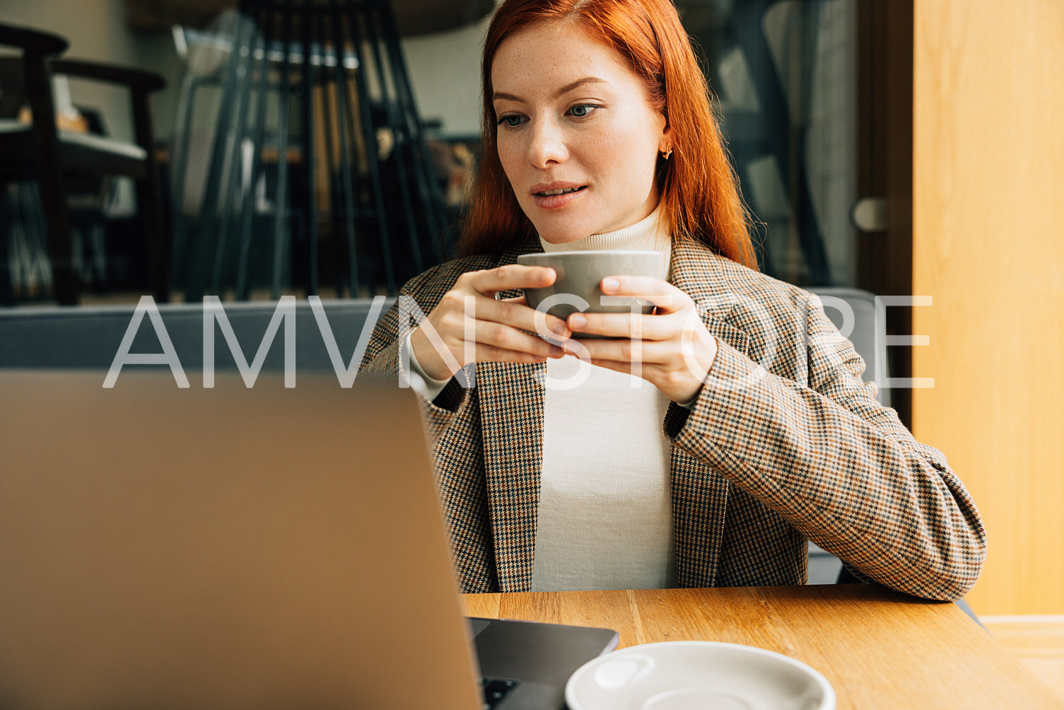 Beautiful woman with ginger hair holding a cup of coffee with two hands looking at a laptop