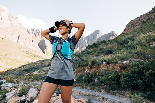 Happy woman hiker with hands on cap. Smiling female enjoying the view while standing in valley and looking at the mountains.